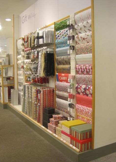 Flat sheets of wrapping paper on the right over dowel rods, a base for stacking gift boxes, acrylic organizers for rolled gift wrap ... plus hooks for tissue paper and gift totes above. Viola! Gift Wrapping Shop Interior, Paper Store Display, Gift Wrap Display Retail, Gift Wrapping Room Ideas, Retail Gift Wrapping Station, Stationary Store Design, Wrapping Paper Display, Gift Wrapping Room, Store Tissue Paper