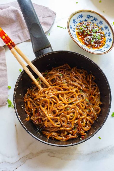 A pan filled with garlic chili oil noodles mixed in a rich, dark sauce is presented on a white marble surface. Wooden chopsticks rest on the edge of the pan. A small blue and white dish containing spicy chili sauce with green garnishes is placed beside the pan. Garlic Chili Oil Noodles, Spicy Chili Oil, Chili Oil Noodles, Spicy Asian Noodles, Garlic Chili Oil, Spicy Chili Sauce, Oil Noodles, Pan Fried Noodles, Chopsticks Rest