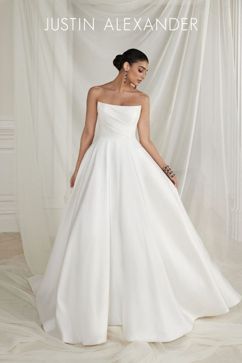 A clean and crisp beauty, this A-line wedding dress is a fashionable take on the minimalistic trend. This luxurious Mikado A-line features a stylized strapless neckline that is accented with asymmetric pleating. A matching bow on the train adds a unique design detail that loops onto the natural waist to instantly create a perfect bustle. The style is finished with pockets and Mikado covered buttons to the end of the chapel length train. Wedding Gown Gallery, Justin Alexander Bridal, Blue By Enzoani, Justin Alexander Wedding Dress, Debutante Ball, Aline Wedding Dress, Bridal Elegance, Blush Bridal, Justin Alexander