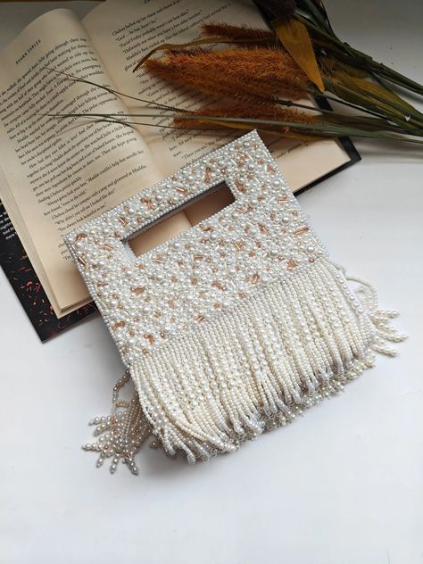 beaded handbag with pearl-like beads and fringe detailing, placed in front of an open book with visible text on a white surface. The cozy reading atmosphere is enhanced by the presence of dried yellow flowers and another partially visible book in the background. Seed Bead Purse, Beaded Square, Beaded Shoulder Bag, Beaded Clutch Purse, Wedding Bags, Fringe Purse, Beaded Bag, Beaded Handbag, Evening Purse