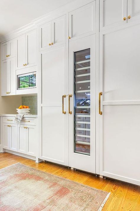 A white wood paneled refrigerator door is accented with an aged brass pull and positioned beneath white shaker cabinets and beside a custom tall glass front wine fridge. Kitchen With White Appliances, Tall Wine Fridge, Wet Bar Cabinets, Paneled Refrigerator, Gorgeous White Kitchen, Gray Shaker Cabinets, White Wood Paneling, Fridge Design, Black Refrigerator