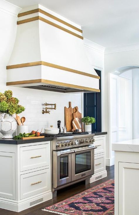 A gorgeous white and gold French range hood is mounted to white subway backsplash tiles over an antique brass pot filler and a stainless steel dual range flanked by white drawers donning brass pulls and a black countertop. Gold Range Hood, White Kitchen Hood, White Herringbone Tile, Kitchen Hood Design, Top Kitchen Trends, White Shaker Cabinets, White Kitchen Island, Cabinet Kitchen, Top Kitchen