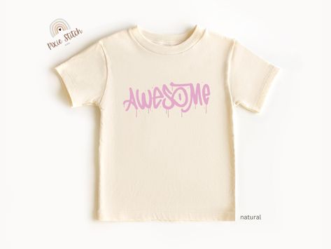 A cream-colored T-shirt with the word “Awesome” in bold, dripping pink letters, resembling a graffiti style. The font gives a playful and energetic vibe. Toddler Tshirt Ideas, Graffiti Rose, Cool Street Art, Pink Graffiti, Vibrant Personality, Chemise Rose, Graffiti Designs, Cool Graphic Tees, Graffiti Styles