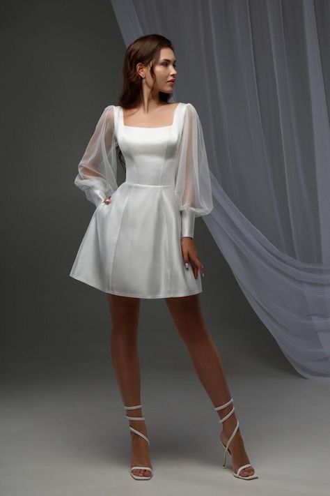 Dive into elegance with our Minimalist White Mini Dress, perfect for any cocktail event or special occasion! Its off-white satin fabric shimmers subtly, making you the center of attention.  - Square neckline & sheer lantern sleeves: A blend of sophistication and charm. - Corset top & natural waistline: Flaunts your figure while the mini length adds a playful twist.  - Pockets & lace-up back: Combine functionality with style. Ideal for summer weddings as a bridal mini dress or an engagement party White Bridal Cocktail Dress, White Wedding Cocktail Dress, Short Balloon Sleeve Wedding Dress, Short Wedding Dress Retro, Cocktail Bridal Dress, Long Sleeve Mini Dress White, Mini Wedding Dress With Sleeves, Skater Wedding Dress, Short White Vintage Dress