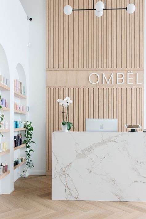 The clean-lined and nature-based design reflects the salon's committment to sustainability in the beauty industry.  Hallmarks of the design are the fluted feature wall, arched product display shelving in white oak, herringbone floors, camel-toned styling chairs, half-moon satin brass hardware, and reception desk wrapped in Dekton Entzo by Cosentino. White And Brass Desk, Natural Retail Store Design, Scandinavian Clinic Interior Design, Boho Reception Desk Design, Day Spa Reception Area, Beauty Salon Feature Wall, Coastal Reception Desk, Accent Wall Behind Reception Desk, Product Display Wall Design