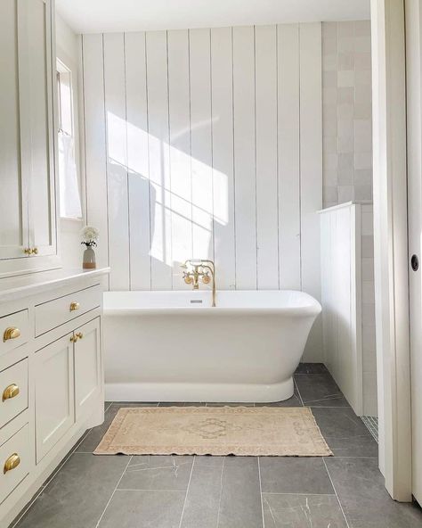 White shiplap paneling is placed vertically around a white freestanding bathtub with gold faucet. Gray floor tile extends across to a tiled shower area with a half wall. White cabinets with gold hardware provide storage in this bathroom. Tile Flooring Bathroom, Dark Grey Tile Bathroom, Dark Floor Bathroom, Mudroom Tile, Tudor Renovation, Dark Tile Floors, Marble Countertops Bathroom, Grey Bathroom Floor, Boy Bathroom