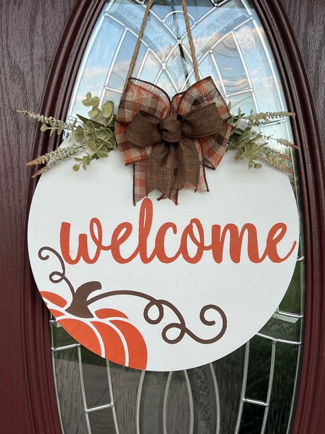 Add a touch of fall to your front door with this "Welcome" round wooden door hanger featuring an adorable pumpkin!  Comes in your choice of two sizes, 15" or 18" round.  Complete and the beautiful bow,  greenery and attached rope for hanging. Wood round details: -Choose your size- 15 inches round (as shown is pictures) or 18 inches round -Greenery and bow are included and attached with special adhesive and staples -Rope is attached on the back for easy hanging -Wood is sealed for protection but Diy Thanksgiving Door Hanger, Wood Rounds Door Hanger Fall, Fall Door Hangers Wooden Diy, Diy Wooden Circle Sign, Diy Wood Wreaths For Front Door, Wood Circle Craft Ideas, Circle Wood Door Sign, Fall Door Hangers Diy, Fall Welcome Sign Front Door