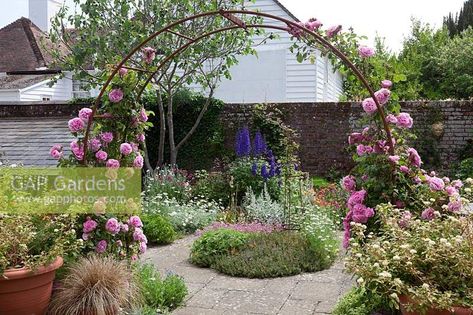 A rusty metal rose arch festooned with Rosa 'Gertrude Jekyll' - Climbing Rose. Set in small courtyard with flower beds, plants: Delphinium 'Black Knight', Geum ' Prinses Juliana', Penstemon 'Garnet', Nigella damascena 'Miss Jekyll' - Love in the Mist, Anthemis, Lychnis coronaria, Artemisia ludoviciana 'Valerie Finnis' Penstemon Garnet, Valerie Finnis, Artemisia Ludoviciana, Lychnis Coronaria, Love In The Mist, Nigella Damascena, Rose Arch, Gertrude Jekyll, Small Courtyard