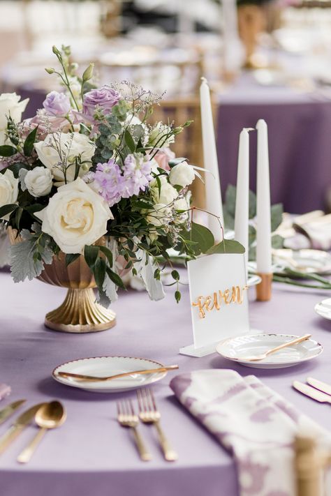 Table Place Setting Decor with Lavender Linens, Gold Silverware, White Taper Candles, and White and Purple Floral Centerpiece Arrangement Light Purple Wedding Table Decor, Lilac Wedding Centrepiece, Purple Table Linens Wedding, Purple And Green Reception Decor, Lavender Place Setting, Sage And Lavender Centerpieces, Lilac And White Decorations, Light Purple Wedding Centerpieces, Lilac And Sage Table Setting