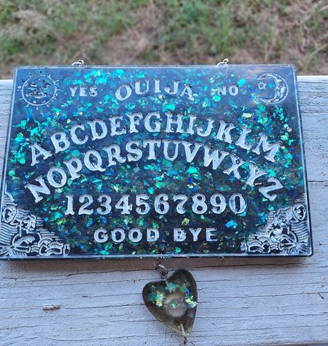 This is a resin cast ouija board full of chunky and sparkly glitter and backed in a dark Grey resin. Symbols are filled with white paint. There is a tiny hanging matching planchet on the bottom with a crystal star hanging below it. The board hangs from a silver chain. Back is not domed. Resin Glitter, Board Wall, Ouija Board, Store Ideas, Crystal Stars, Wall Board, White Paint, White Paints, Silver Chain