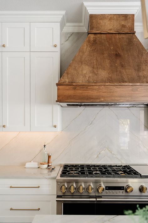 We love the combination of this stone backsplash and wood-stained vent hood. The brass hardware and crisp white cabinets complete this new traditional style kitchen design. Range Next To Doorway, Kitchen Backsplash Behind Hood, White Kitchen Hood Ideas, Stained Vent Hood, Kitchen Vent Hood Ideas Wood, Wooden Vent Hood Ideas, Wood Kitchen Hood, Stove Backsplash Ideas, Wooden Vent Hood