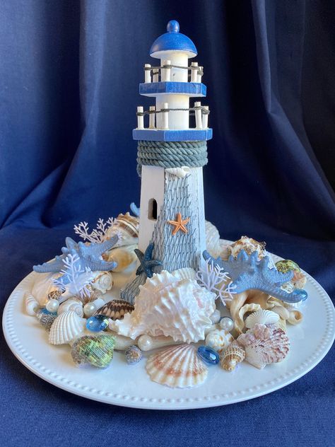 Tall Wood Lighthouse & Shells Nautical Centerpiece w/Slate Blue Starfish, Sand Dollar, Driftwood, Pearls, on White Charger The lighthouses have different designs on the netting, chosen at random. This one of a kind centerpiece is a special vignette to grace your coffee table, dining table, bringing a floral beach feeling to your decor. Or it would be so nice on the entry table to announce to your guests that beach lovers live here. It would make a charming addition to your patio table as well, but please bring back inside after use as it is not weather resistant. The shells in this arrangement are permanently set in place. Please remember that sea shells are delicate and fragile and can also have sharp edges. Use caution with children. While this creation may be used outside, it is not mea Seashell Table Decor Centerpieces, Nautical Centerpiece Ideas, Nautical Centerpieces, Beach Theme Centerpieces, Nautical Centerpiece, Wood Lighthouse, Shell Centerpieces, Beach Themed Crafts, White Charger