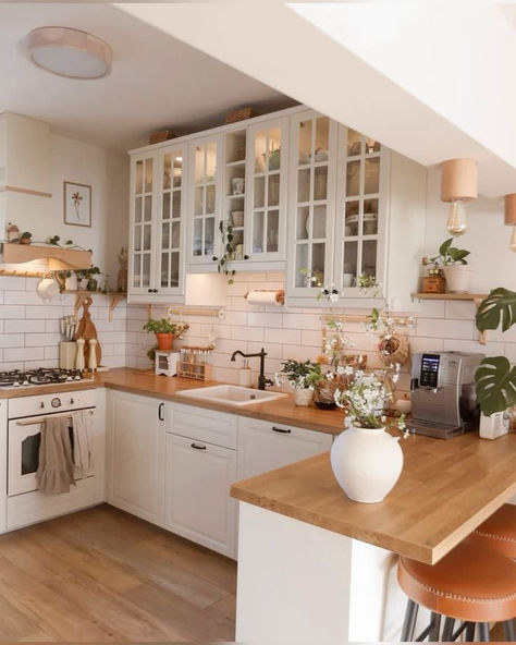 A cozy kitchen with cream cabinets, wooden countertops, glass-paneled cabinets, and modern appliances. Plants and natural light enhance the welcoming atmosphere. Warm Apartment Aesthetic, Warm Apartment, Kitchen Cream, Den Decor, Wooden Countertops, Organized Kitchen, Perfectly Organized, Apartment Aesthetic, Cream Tones