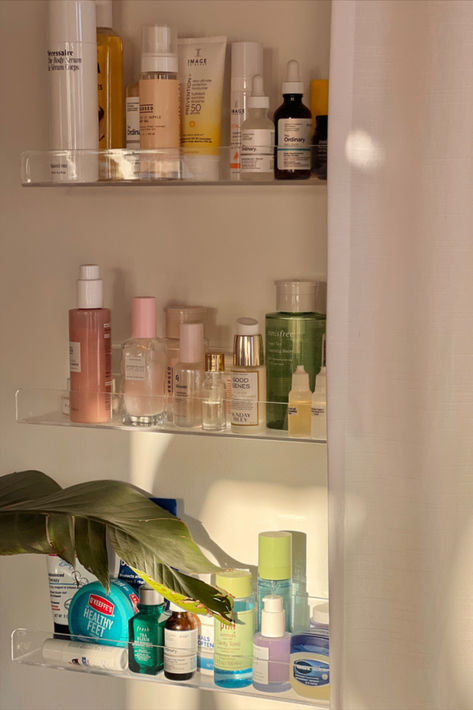 A set of clear acrylic shelves mounted on a wall, displaying an array of skincare products in a neat and organized manner. The shelves hold various serums, lotions, and creams, highlighting the beauty of clear storage solutions. Skincare Wall Shelf, Kids Book Display, Book Display Shelves, Skincare Display, Skincare Shelf, Clear Shelves, Organization Skincare, Acrylic Shelves, Book Display Shelf