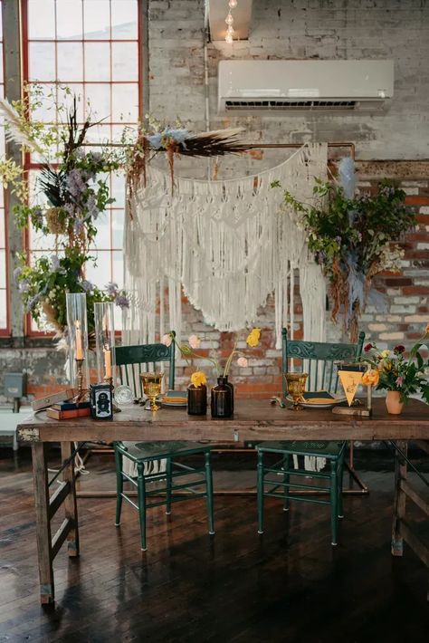 A vintage-inspired sweetheart table with low, minimalistic centerpieces, candles, colored goblets, books and a camp flag to go with the Wes Anderson-inspired wedding reception, all in front of a macrame backdrop on a wood arch and verdant floral arrangements. Sweetheart Chairs Wedding, Minimalistic Centerpieces, Colored Goblets, Centerpieces Candles, Camp Flag, Macrame Wedding Arch, Geneva New York, Wedding Glassware, Table Halloween