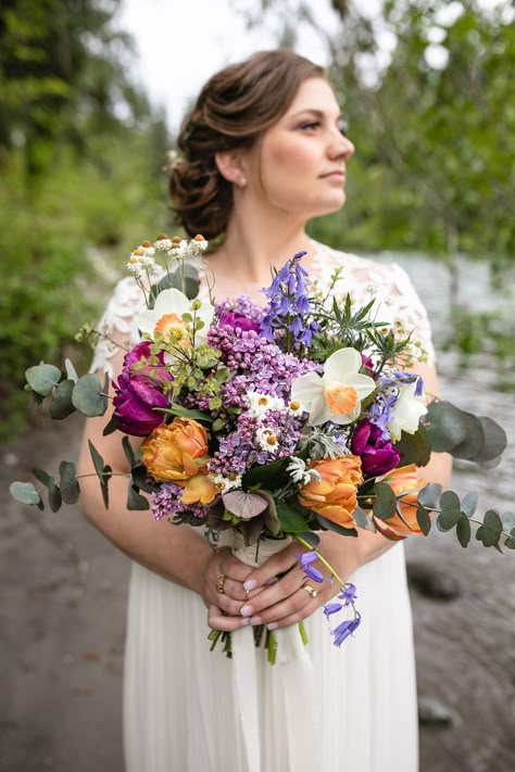 Bride standing by the river with lush bouquet of purple lilacs, white and orange daffodils, orange tulips, and eucalyptus foliage. Bouquet With Lilacs, Wedding Bouquet Lilac, Bouquet Eucalyptus, Lilac Bridal Bouquet, Purple Orange And Green Wedding Colors, Lilac And Orange Wedding Flowers, Peach Purple Wedding, Purple And Peach Flowers, Purple And Yellow Bridal Bouquet