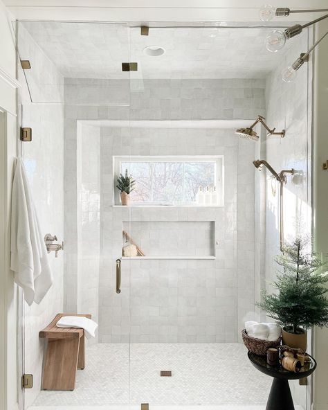 A wood bench is placed behind the glass panels of a shower with a window. The window is surrounded by white marble tile and a rectangular shower niche. Nickel shower heads are anchored to one wall. Subtle nature decor accessorizes a modern black table. Honey Marble Bathroom, Zia Tile Bathroom, Vanilla Bathroom, Bathroom Gold Fixtures, Dr Bathroom, Gold And White Bathroom, Shower Windows Ideas, White And Gold Bathroom Decor, Cali House