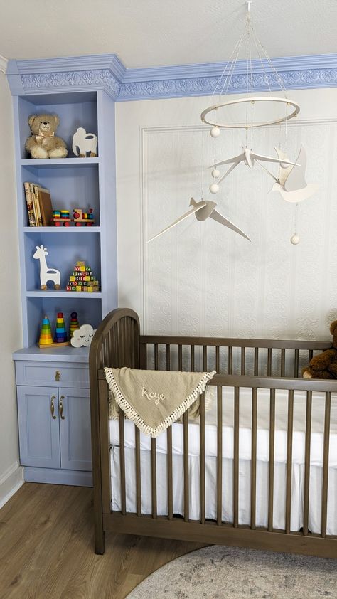 The left corner of a nursery. In the corner there are periwinkle floor to ceiling built in shelves. The crown molding connects with the shelves and is also periwinkle. The walls are covered in textured wallpaper. In front of the wall is a brown crib with a neutral rug beneath it. Hanging over the crib is an off-white mobile with birds and spheres on it. Built In Crib, Molding Wallpaper, Nursery Room Diy, Shelves Nursery, Nursery Design Neutral, Nursery Room Inspiration, Diy Nursery, Built In Shelves, Crown Molding