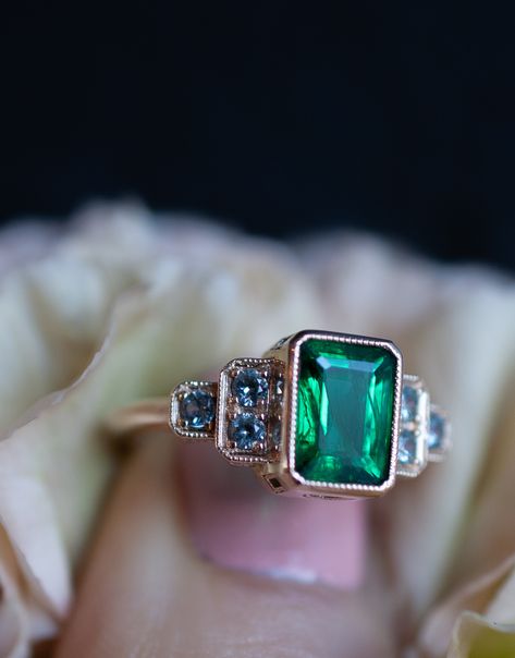 An emerald and sapphire engagement ring with a bezel set emerald cut green emerald center stone set in contrasting yellow gold. The unique custom ring was made to stand out as a bold, Art Deco inspired non-diamond engagement ring. A classic setting has vintage style milgrain around all stones. Six unique pale blue grey Montana sapphires frame the vivid emerald in an unexpected flourish. #gemstonengagementring #emeraldring #uniqueengagementring Emerald Antique Engagement Ring, Emerald And Sapphire Engagement Ring, Emerald Sapphire Engagement Ring, Sapphire And Emerald Engagement Ring, Unique Emerald Engagement Ring, Art Deco Emerald Engagement Ring, Emerald Art Deco Engagement Ring, Sapphire And Emerald Ring, Emerald Art Deco Ring