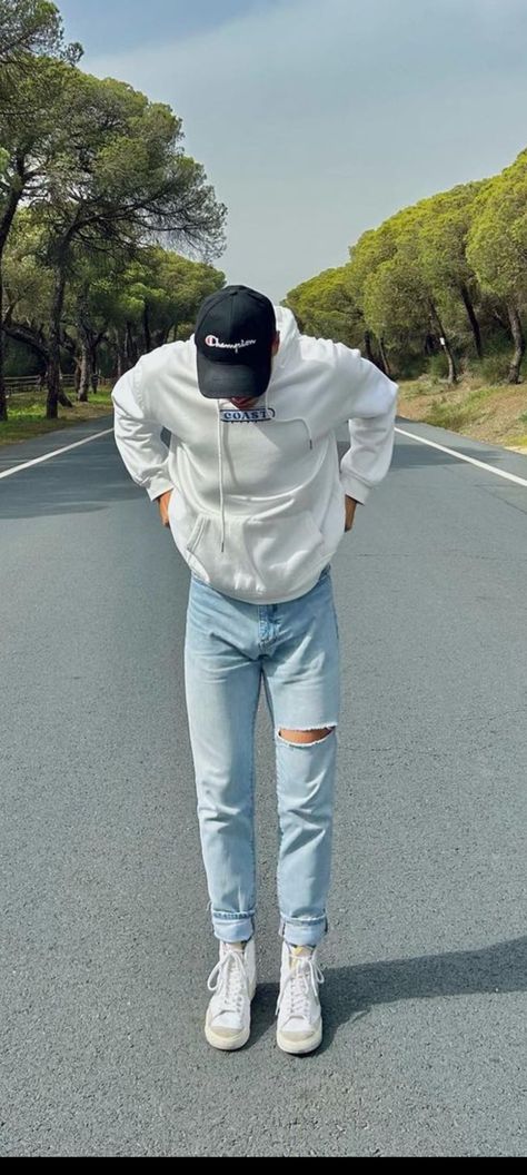 A white man is on a road wearing a black cap, a white shirt, ripped jeans and white shoes. There are trees in the sides of the road. Light Wash Denim Outfit Men, Lightwash Jean Outfit Men, Light Denim Pants Outfit, Mens Light Blue Jeans Outfit, Light Blue Jeans Outfit Men Casual, Light Blue Denim Jeans Outfit Men, White Shirt Blue Jeans Men, Sky Blue Jeans Outfit Men, Light Jeans Outfit Men