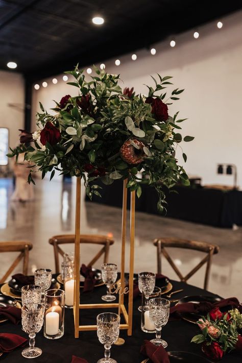 Tall centerpieces on every other dining table adds interest to the room.  These gold harlow stands supporting the floral arrangement made of deep red roses and king proteas sparkle against the dramatic black table linens. Dark And Moody Centerpiece, Burgundy And Black Floral Arrangements, Tall Moody Wedding Centerpieces, Burgandy Black Rose Gold Wedding Decor, Eucalyptus Table Setting, Winery Wedding Centerpieces, Black Table Linens Wedding, Black Tablecloth Wedding, Protea Centerpiece