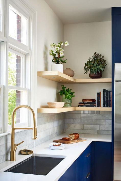 Brass fixtures are a warm glow over the sink, the gold color nicely complementing the floating shelves' wood. Marble tile layers in depth on the kitchen's backsplash. Navy Blue And White Kitchen, Village Family, Blue And White Kitchen, Family Dinner Table, Mediterranean Kitchen, Cozy Breakfast Nook, Chinoiserie Decorating, Transitional Living, Blue Cabinets