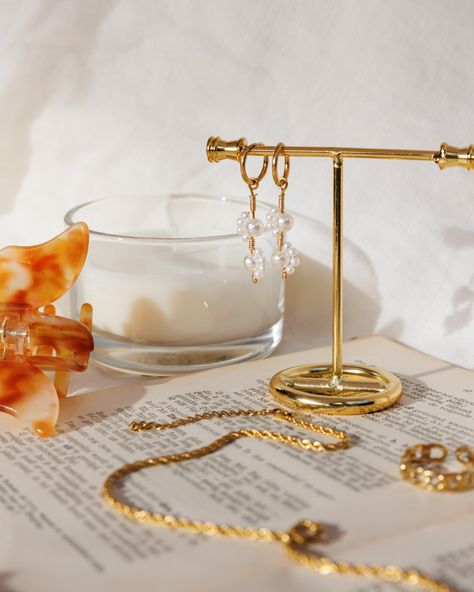 Gold pearl earrings hanging on display, on top of book with other gold jewelry & a candle in the background Asthetic Jwellery Photography, Jewlrey Aesthic Photography, Summer Jewelry Photoshoot Ideas, Jewelry Product Photography Hacks, Necklace Photography Ideas Model, Delicate Jewelry Aesthetic, Product Photography Earrings, Earrings Product Photography, Earing Photography Ideas