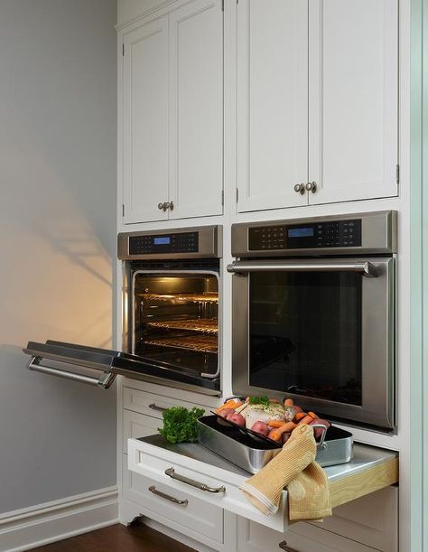 Under white shaker cabinets, side-by-side stainless steel wall ovens are mounted above pull out drawers donning polished nickel pulls and finished with pull out stainless steel countertops. Kitchen Corner Stove, Stove Wall, Corner Stove, Oven Cabinet, Oven Design, Wall Ovens, White Shaker Cabinets, Stainless Steel Oven, Transitional Kitchen