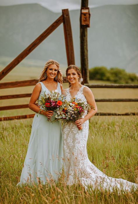 A wildflower inspired wedding in June near the bighorn mountains in sheridan wy with bouquets that are brightly colors and feature asters, wax flower, gerbera daisies and colors in white, orange, pink and purple.  bridemaid wearing a long dress in pale blue green and standing with a bride wearing a v-neck dress with lace overlay in a field of grass with mountains and fenceline in the background Fall Wildflower Wedding Bridesmaids, Green Bridesmaid Dresses Wildflowers, Sage Green Bridesmaid Dresses With Wildflowers, Sage Green Dresses, Texas Wildflowers Bouquet, Flattering Wedding Dress, Wildflower Bouquets, Wyoming Wedding, Wildflowers And Wild Horses