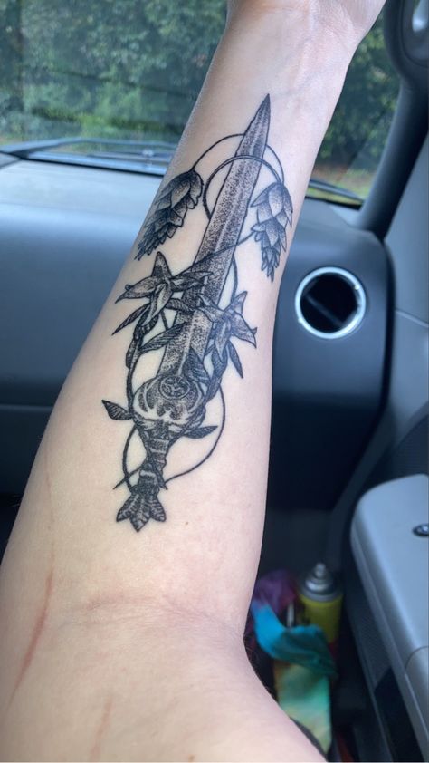 A black and grey tattoo on the inner forearm of a sword from the Skyrim video game with a bird design on the hilt and flowers surrounding it. Skyrim Nocturnal Tattoo, Subtle Skyrim Tattoo, Skyrim Nightingale Tattoo, Paarthurnax Skyrim Tattoo, Nocturnal Skyrim, Skyrim Tattoo Ideas, Skyrim Swords, Video Game Tattoo Ideas, Nightingale Tattoo