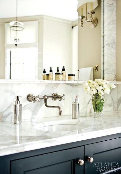 Black bathroom vanity with satin nickel knobs is accented with gray and white marble countertops holding an oval sink completed with a vintage cross handle faucet mounted on a marble backsplash under a marble shelf and a marble famed vanity mirror. Alternative Bathroom, Vanity Backsplash, Classic Bathroom Design, Black Vanity Bathroom, Marble Shelf, Bathroom Storage Solutions, Medicine Cabinets, Marble Backsplash, Wall Mount Faucet