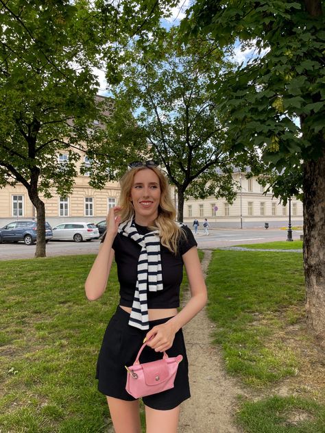 A girl wearing a black top with black shorts and a black and white striped top tied around shoulders, with a small pink handbag from the brand longchamp, in the background there is a small park with green trees and some European buildings. Small Longchamp Bag Outfit, Pink Longchamp Bag Outfit, Mini Longchamp Bag Outfit, Longchamp Mini Pouch, Mini Longchamp Bag, Pink Longchamp Bag, Longchamp Pouch, Longchamp Bag Outfit, Pink Bags Outfit