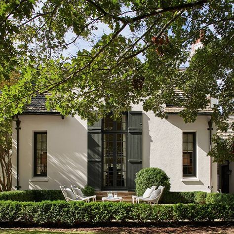 A creamy white-painted stucco home with tall center glass doors and dark green door shutters and trim, and steps to patio furniture surrounded by a green hedge and trees. Steps To Patio, Dark Green Door, Tudor Exterior Paint, Stucco Exterior Colors, Painted Stucco, White Stucco House, Benjamin Moore Exterior Paint, Door Shutters, Green House Exterior