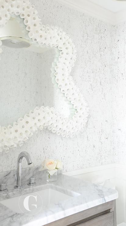 Gorgeous white and gray beach bathroom boasts a gray wash washstand topped with a carrera marble countertop fitted with an undermount sink and polished nickel gooseneck faucet mounted in front of a carrera marble backsplash fixed beneath a white coral framed mirror mounted to a white and gray wallpapered wall. Carrera Marble Backsplash, Half Bath Wallpaper, Coral Mirror, Octopus Bathroom, Beachy Bathroom, Beach Bathroom, Dream Beach Houses, Carrera Marble, Coastal Bathrooms