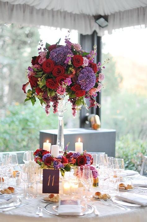 Pink and red roses, purple peonies, burgundy Calla lilies and bunches of grapes top clear pedestals in these elegant centerpieces. Wedding Centerpieces Elegant Romantic, Wedding Centerpieces Elegant, Purple White Wedding, Pink And Red Roses, Roses Purple, Elegant Wedding Centerpiece, Purple Table, Wedding Color Combos, Purple Wedding Bouquets