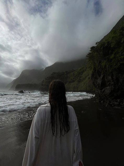 A picture of a girl on the Seixal beach on Madeira. Her wet hair from the sea is falling down onto her white shirt. She is facing the sea, not the camera. The sky is cloudy without the sun brightening it up, so the colours seem dark. Black Sand Beach Photo Ideas, Black Sand Beach Aesthetic, Madeira Outfit Ideas, Seixal Beach Madeira, Cloudy Beach Photoshoot, Cloudy Beach Aesthetic, Madeira Portugal Aesthetic, Wet Hair Aesthetic, Cloudy Photoshoot