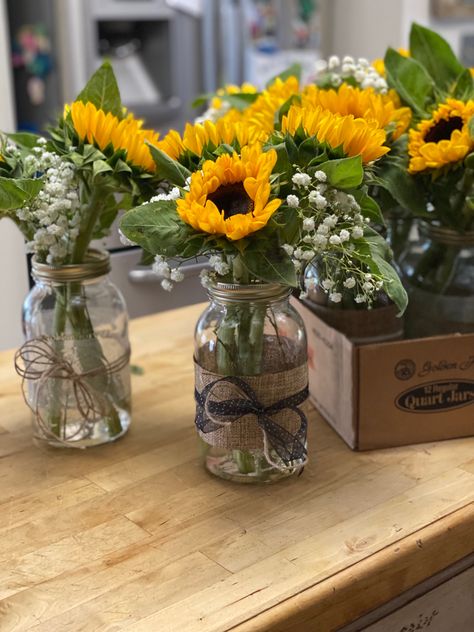 Sunflower centerpieces for our daughter’s graduation party. Mason jars from Walmart, sunflowers and baby’s breath from local grocery store, and ribbons and twine from Michael’s. I used double-side tape to attach the wide ribbon to the jars then tied the smaller ribbon around that. Add about 2-3 inches of water and you’re done. Sunflower Mason Jar Centerpieces, June Wedding Colors, Fairy Lights Wedding, Sunflower Centerpieces, Sunflower Vase, Party Table Centerpieces, Western Party, Wide Mouth Mason Jars, Local Grocery Store