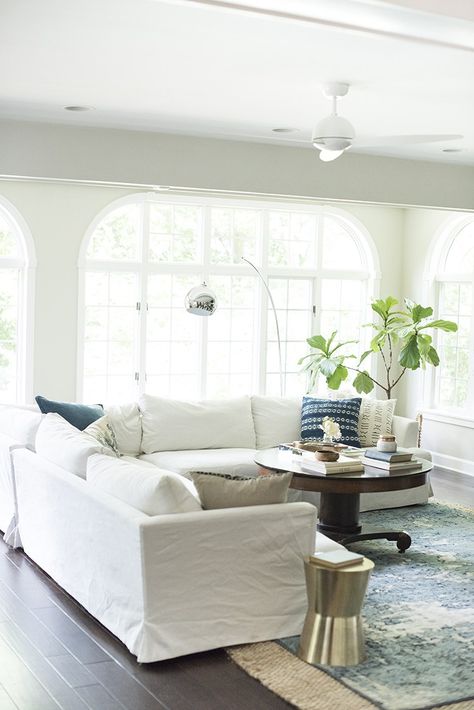 White sectional in boho living room with layered rugs and giant windows Two White Sofas Living Room, White Couch Cover Sofa Slipcovers, White Slip Cover Couch, Slipcover Couch Living Room, White Linen Couch Living Room, White Slip Covered Sofa Living Room, Slip Covered Sofa Living Room, Slip Cover Sofa Living Room, Living Rooms With White Couches