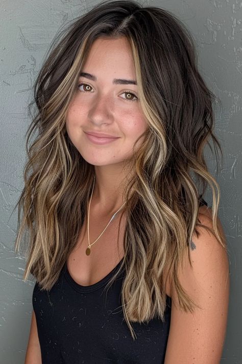 Woman with wavy, highlighted hair smiling against a gray textured wall. Brunette Hair Peekaboo Highlights, Dark Brown Medium Hair With Highlights, Brown Hair With Face Highlights, Brown Fall Hair With Highlights, Cute Dark Hair Ideas, Accent Balayage Brunettes, Dramatic Dimensional Hair Color, Dark Brunette Highlights Lowlights, Hair Dark With Highlights