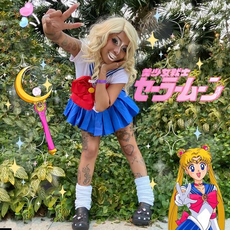 A mixed black girl is posed leaning towards the camera making a peace sign with her right hand. She is wearing gyaru make up, white eyeshadow, white lipstick, blonde curly hair, has assorted tattoos on her legs and arms,wearing a sailor moon cosplay, with slouched socks and black crocs.  She is standing in front of foliage. The photo stickers are the Japanese Sailor moon logo and the sailor moon stick. End description. Kuronba Gyaru, Manba Gyaru Outfits, Gyaru Halloween, Banba Gyaru, Gyaru Subcultures, Black Sailor Moon, Manba Gyaru, Kuro Gyaru, Black Gyaru