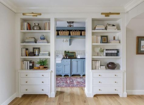 Brass picture lights illuminate white built-in bookshelves fitted over white drawers donning brass hardware and flanking a laundry room doorway. Gold Grasscloth Wallpaper, Mirrored Kitchen Cabinet, Cottage Entrance, Styled Bookshelves, Brass Picture Light, Bookshelf Lighting, Light Gray Cabinets, Bookcase Lighting, Picture Lights