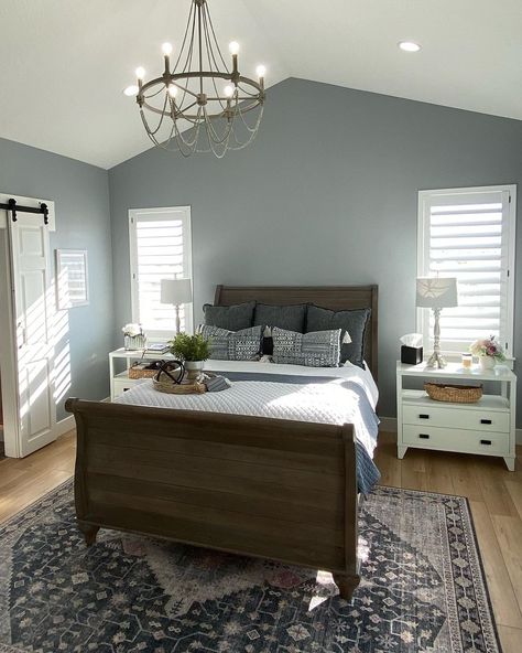 The primary bedroom in this photograph is home to a full-size bedframe made of deep walnut between two shuttered windows and white nightstands. A gray beaded chandelier extends from a pitched ceiling, accentuated by blue-gray walls. To help spotlight the blue undertones of the paint, the designer incorporated denim-colored pillows and a faded Persian rug to line the area underneath the bed, an easy complementary piece to anything with gray undertones. Dark Headboard With White Nightstands, Accent Wall Bedroom Blue Grey, Blue Grey Wood Bedroom, Blue Gray Walls Bedroom, Grey Fabric Bed Contrast Walls, Grey And Blue Bedroom Master Accent Walls, Gray Blue Bedroom Ideas, Blue Grey Bedroom Walls, Bedroom Gray Walls