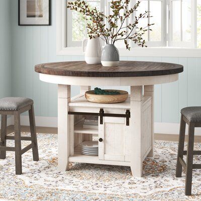 This dining table is made from reclaimed pine wood, which gives it a rustic, coastal farmhouse look. The tabletop has a brown finish that lets the natural wood grain shine through, while its base has a vintage white tone with weathered and distressed details. We love how this table's pedestal base doubles as extra storage. There's a top shelf, a small cabinet, and two cubbies to help get everything off the table when it's time for arts and crafts with the family. Metal hardware detailing rounds Modern Farmhouse Dining Room Table Circle, Small Round Farmhouse Kitchen Table, Small Modern Farmhouse Table, Small Farmhouse Kitchen Table, Small Farmhouse Ideas, Counter Height Kitchen Table, Kitchen Table With Storage, Extendable Pedestal Dining Table, Dining Essentials