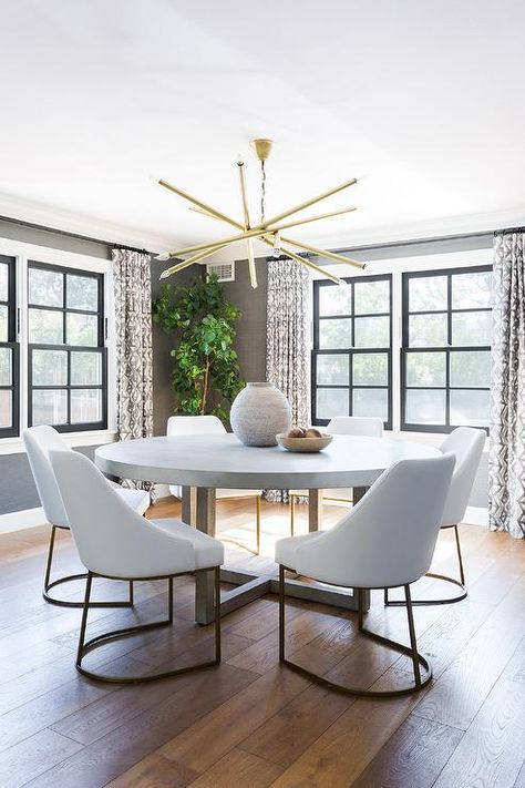 White and gold dining chairs surround a round concrete and wood dining table illuminated by a brass wand chandelier. White Round Dining Table, Gold Dining Chairs, Circular Dining Table, Transitional Dining Room, Gold Dining, Concrete Dining Table, Grey Dining Room, Round Dining Room, White Dining Table