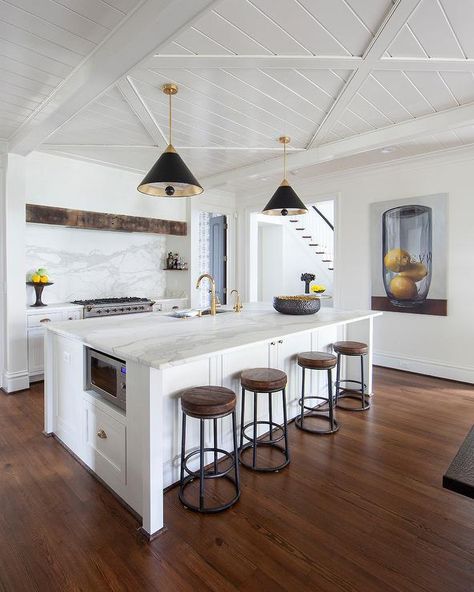 Wood and iron counter stools sit at a honed statuary marble countertop accenting a white kitchen island fitted with an under-counter microwave and a sink with an antique brass faucet. Modern Shaker Style Kitchens, Modern Shaker Style, Luxury White Kitchen, Modern Gold Chandelier, Wood Sink, Metallic Frame, White Shaker Cabinets, White Kitchen Island, Wood Kitchen Island