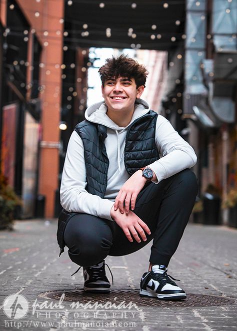 A high school senior boy with curly hair crouches down in an Detroit alleyway decorated with string lights. He is wearing a white hoodie, black puffer vest, black pants, and black and white sneakers. He is smiling and looking away from the camera for his downtown Detroit senior pictures session. The image is by Paul Manoian Photography. Sneaker Senior Pictures, Robotics Senior Pictures, Senior Picture Poses For Guys, Chicago Senior Pictures, Golf Poses, Ballet Senior Pictures, Athena Lee, Boy With Curly Hair, Male Posing