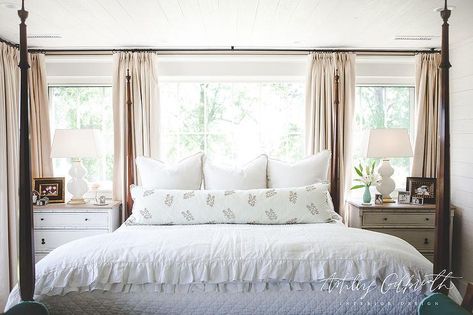 A four poster bed ensues comfort dressed with white pleated bedding, a long white and gray lumbar pillow and a set of three accents pillows in front of large bedroom windows embellished with beige curtains. Bedroom Curtains Behind Bed, Window Behind Bed, Ashley Gilbreath, Ruffled Duvet, Curtains Behind Bed, Cottage Bedrooms, Arch Angel, Bedroom Wall Decor Ideas, Headboard With Shelves