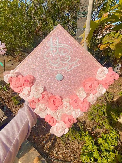 glitter background with white handwritten arabic lettering and paper flowers high school and college senior year graduation cap/hat inspiration, made with acrylic paint Arabic Graduation Cap, Islamic Grad Cap Ideas, Muslim Grad Cap Ideas, Graduation Cap Designs Arabic, Graduation Cap Designs In Arabic, Muslim Graduation Cap, College Senior Year, Flower Grad Cap, Pink Rhinestone Grad Cap