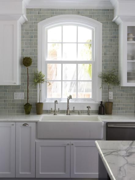 A ceramic farmhouse sink with a brushed-nickel faucet anchors this serene transitional kitchen. Countertop topiaries and a green subway-tile backsplash create an earthy backdrop for crisp white cabinetry and an arched window. Farmhouse Kitchen Backsplash, Kitchen Sink Design, Kitchen Backsplash Designs, Subway Tiles, Gorgeous Kitchens, Modern Farmhouse Kitchens, Trendy Kitchen, Kitchen Redo, Decor Minimalist