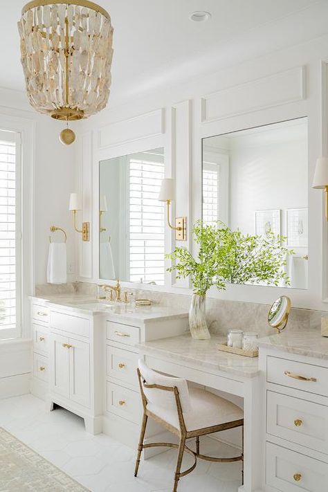 A seashell chandelier hangs over a gray and blue rug accenting a bathroom boasting his and hers white bath vanities donning brass hardware and marble countertops finished with brass faucets mounted under inset mirrors framed by white trim molding. Seashell Chandelier, Blue Bathroom Rugs, Neutral Bathroom Decor, Bamboo Dining Chairs, Brass Faucets, Blue Laundry Rooms, Bathroom Chandelier, Master Bath Renovation, Off White Kitchens